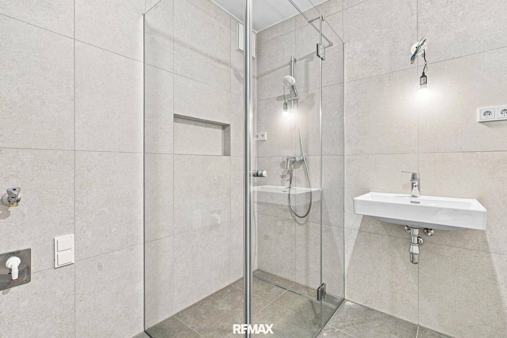 Modernes Badezimmer mit Glasduschkabine, kleinem Waschbecken und hellen Fliesen.