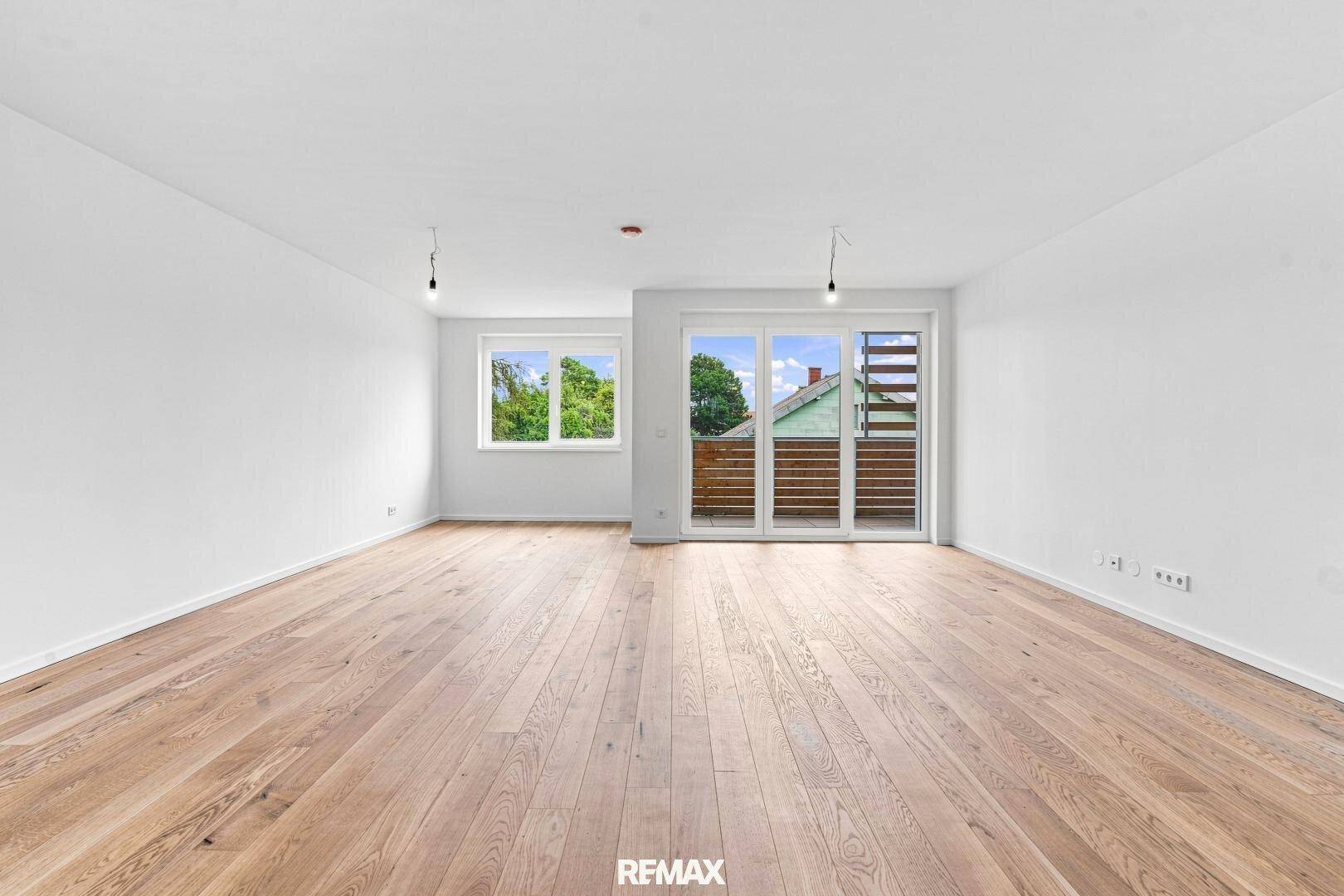 Heller, leerer Wohnbereich mit Holzboden und großen Fenstern zum Balkon.