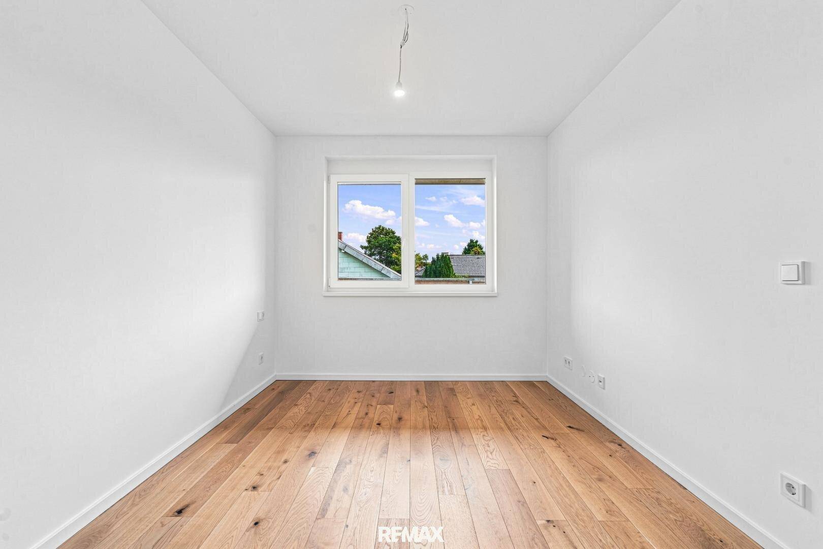 Kleineres, helles Zimmer mit Holzboden und einem Fenster mit Blick ins Grüne.
