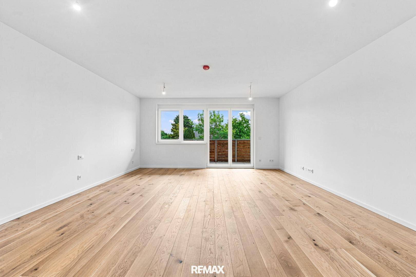 Geräumiger, unmöblierter Raum mit hellem Holzboden und großen Fenstern zum Balkon.