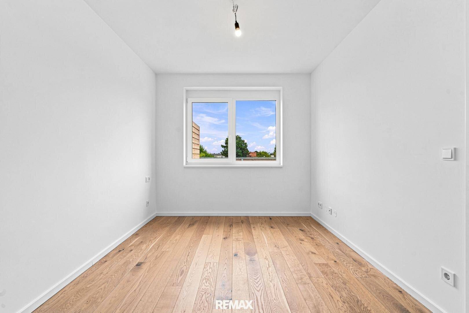 Leeres Schlafzimmer mit Holzboden und einem großen Fenster, das viel natürliches Licht hereinlässt.