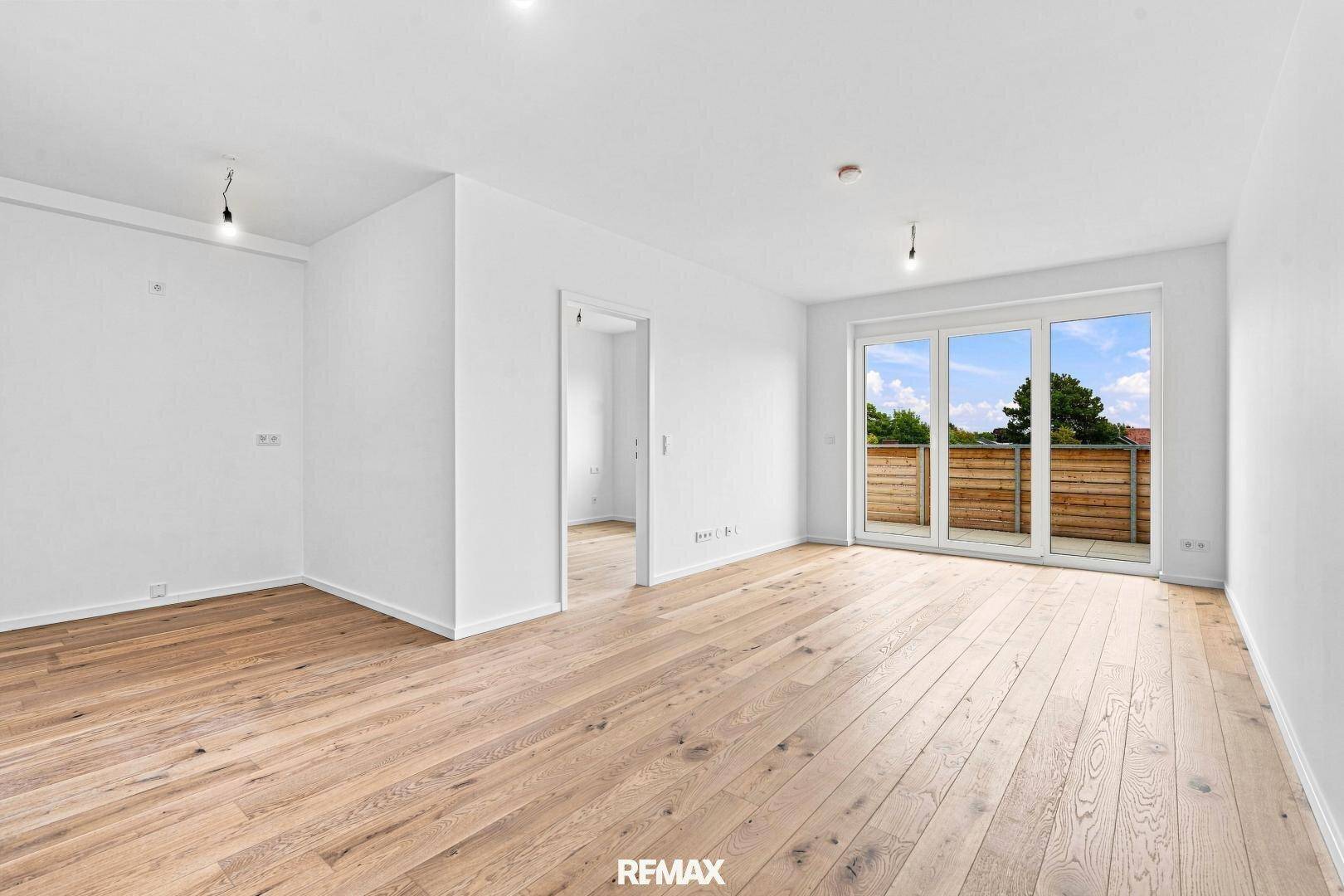 Geräumiges, leeres Wohnzimmer mit hellem Holzboden und großen Fenstern zum Balkon.