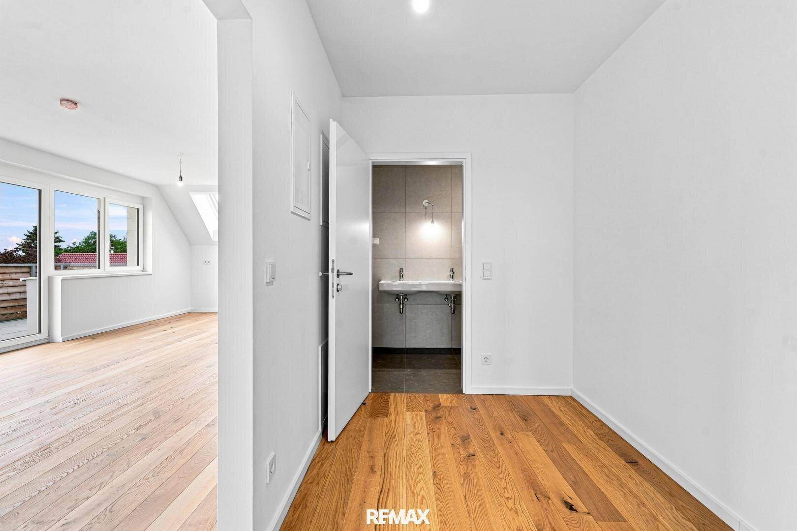 Leeres Zimmer mit Holzboden und offener Tür zu einem modernen Badezimmer mit Doppelwaschbecken.