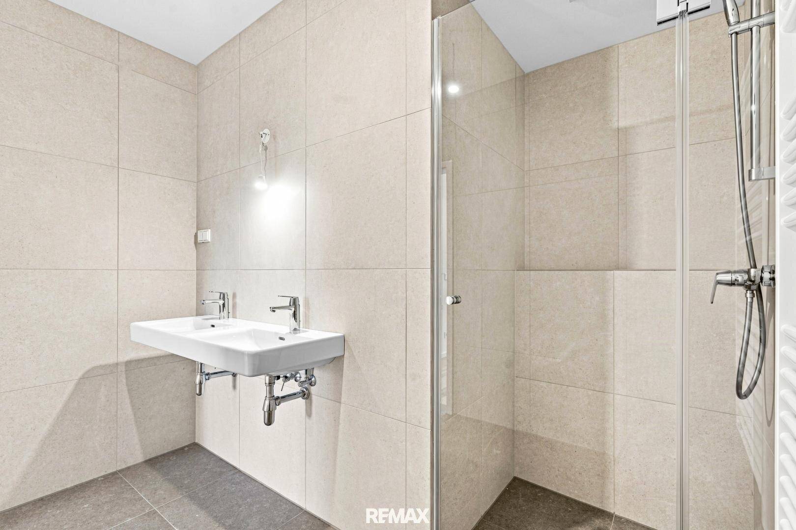 Modernes Badezimmer mit beigefarbenen Fliesen, Doppelwaschbecken und ebenerdiger Dusche mit Glaswand.