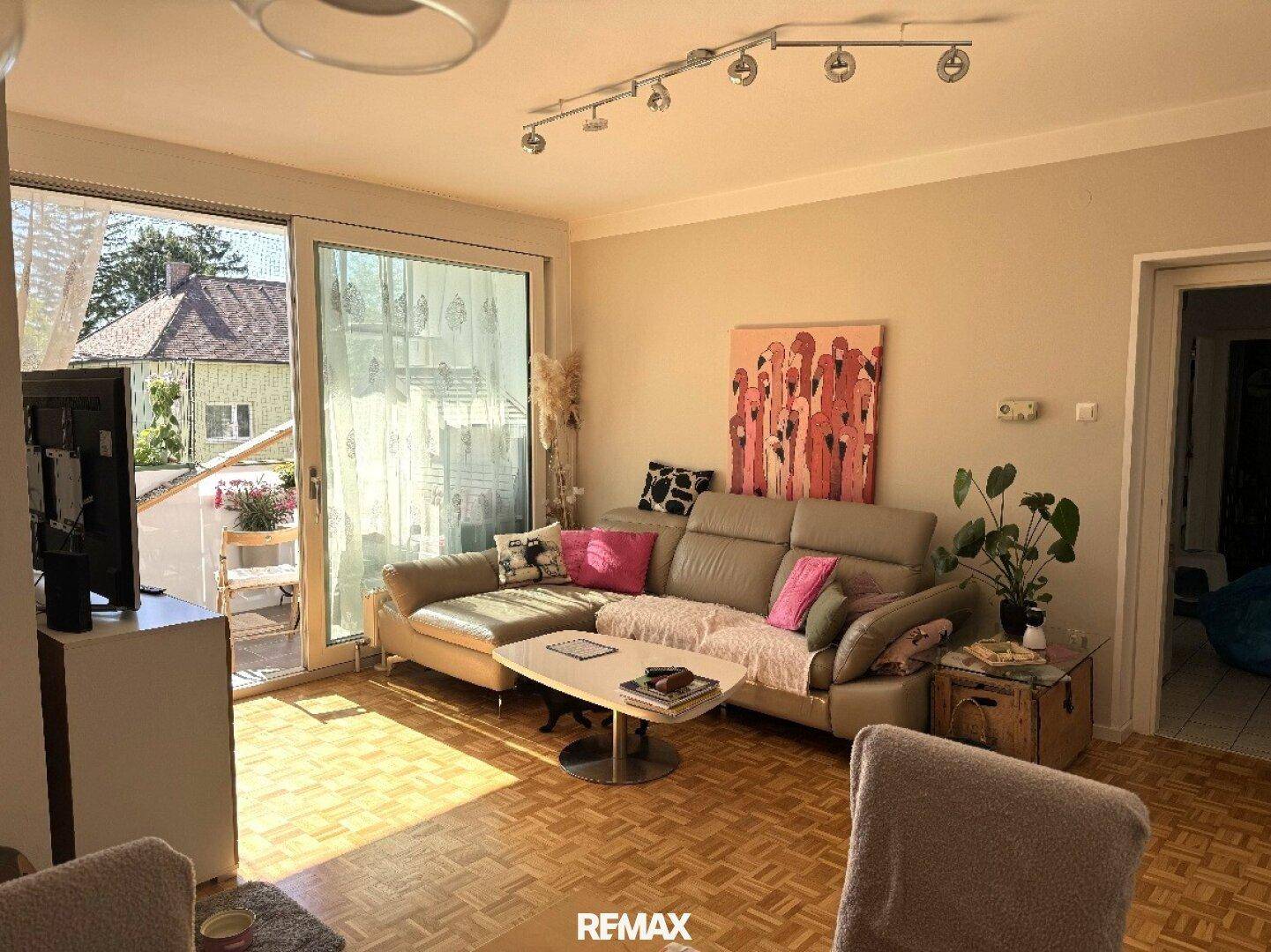 Helles Wohnzimmer mit beigem Sofa, Parkettboden und großem Fenster mit Zugang zum Balkon.