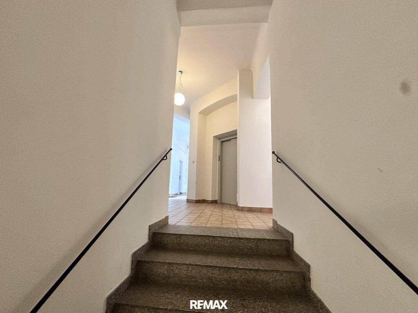 Treppenhaus mit Handlauf und Fliesenboden, das zu einem Flur mit Aufzug führt.
