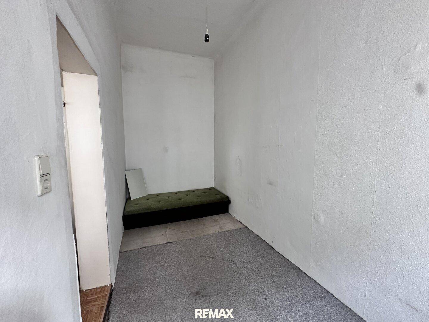 Schmaler Raum mit einfacher Sitzgelegenheit und Teppichboden, nutzbar als Abstellraum.