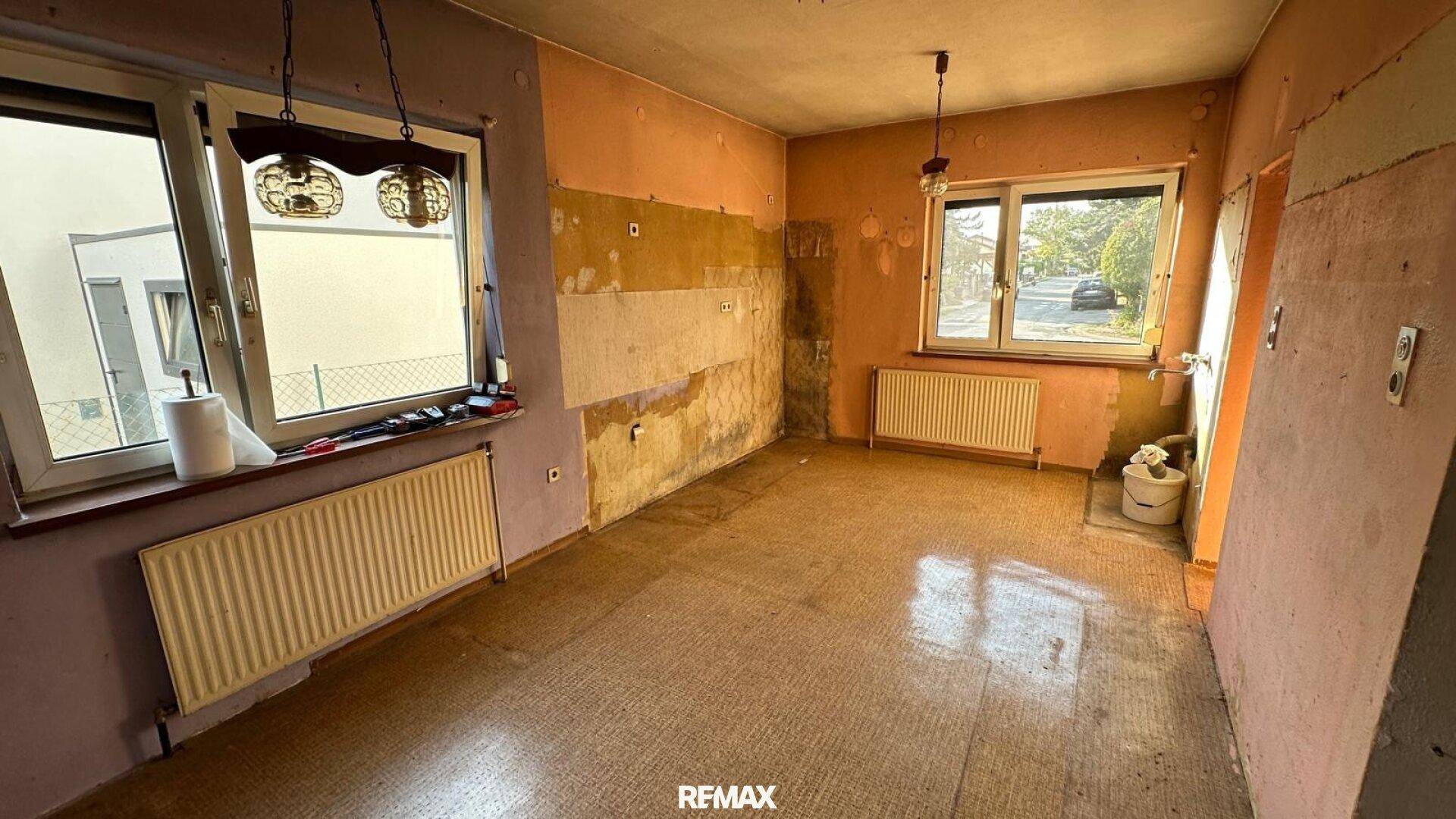 Renovierungsbedürftige Küche mit zwei Fenstern und abgenutzten Wänden.