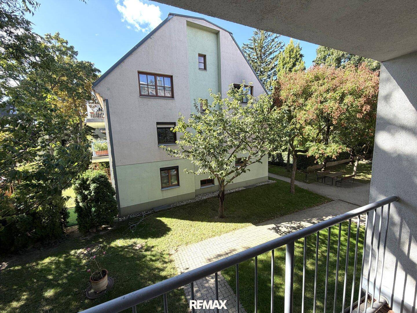 Blick vom Balkon auf die grüne Umgebung und das Mehrfamilienhaus mit Bäumen.
