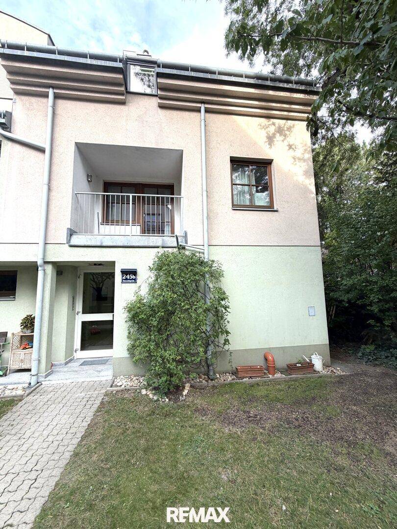 Eingangsbereich des Gebäudes mit Balkon und gepflegter Grünfläche, umgeben von Bäumen.