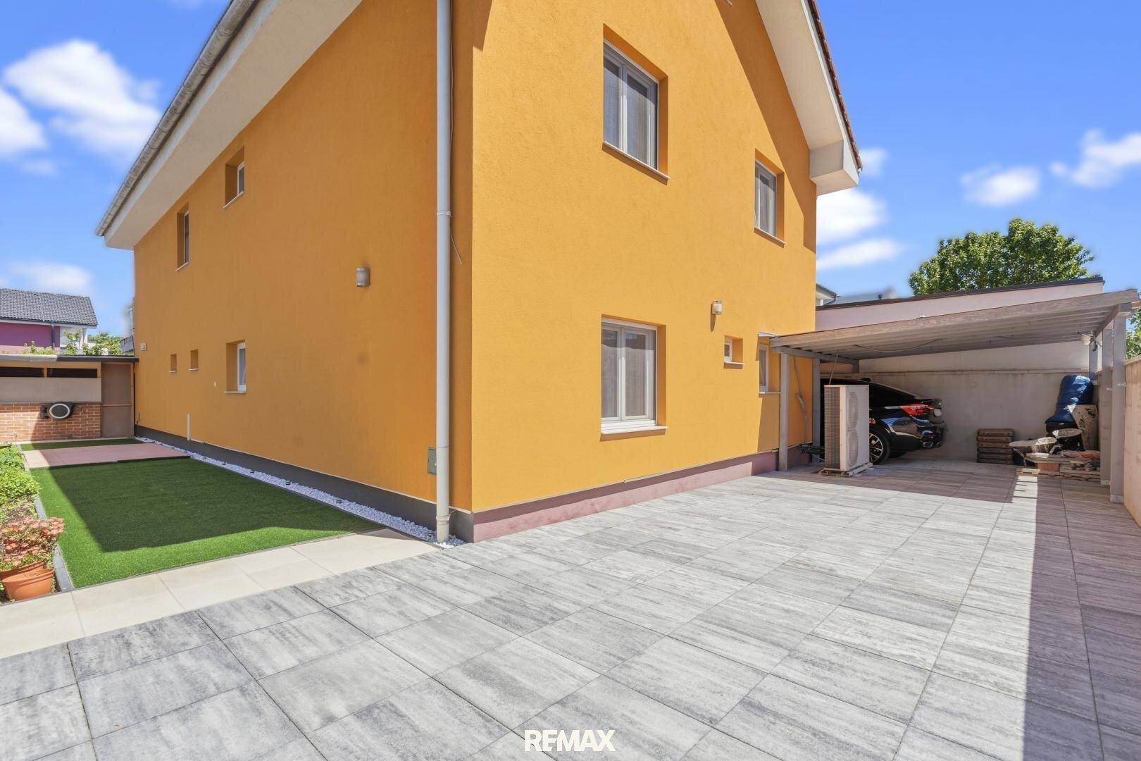 Seitenansicht des Hauses mit gepflasterter Einfahrt, Carport und einem kleinen Rasenbereich.