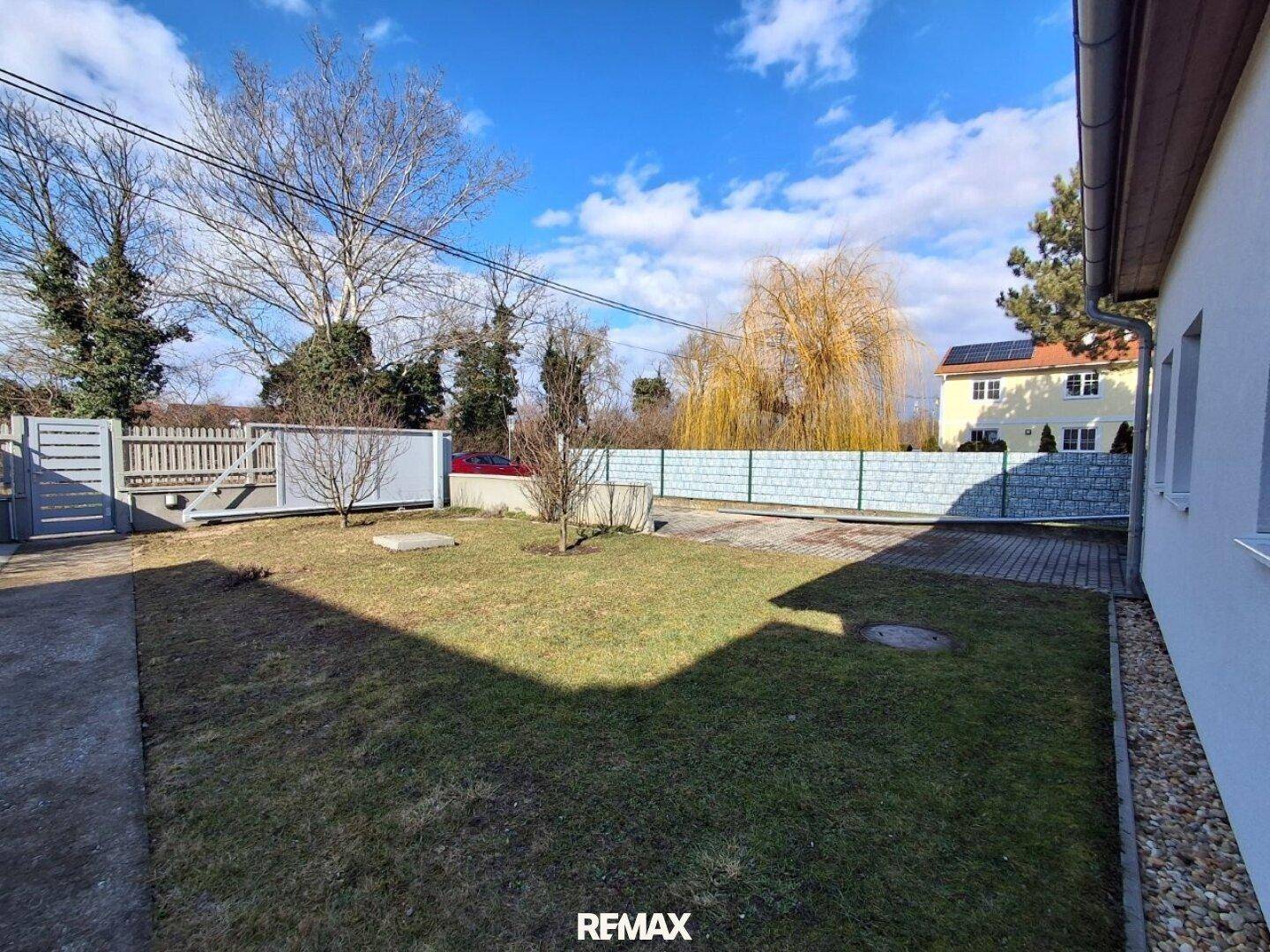 Gartenbereich mit Rasenfläche, gepflastertem Weg und Zaun, umgeben von Bäumen und einem Haus.