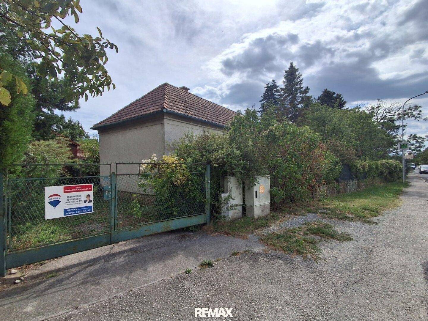 Das Haus von der Straße aus gesehen, mit einem Metalltor und dichtem Bewuchs entlang des Zauns.