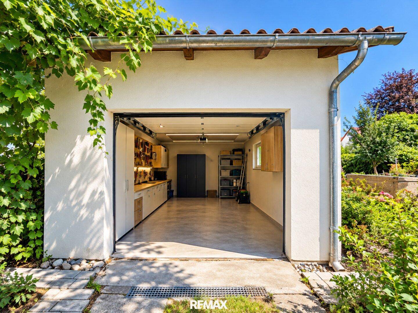 Offene Garage mit Werkbank und Stauraum, umgeben von grüner Bepflanzung.