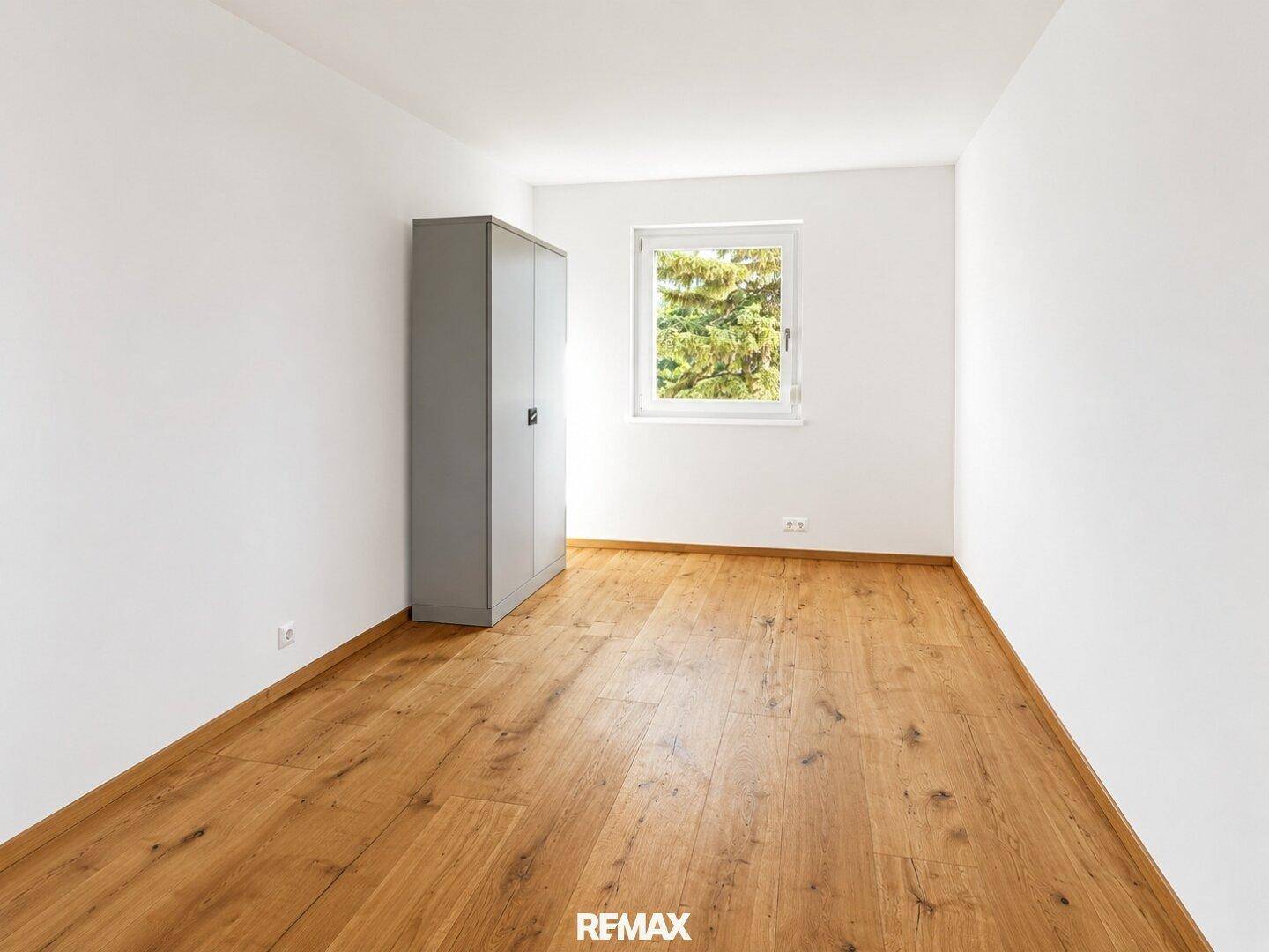 Helles Zimmer mit Holzboden, weiß gestrichenen Wänden und einem Fenster mit Blick ins Grüne.
