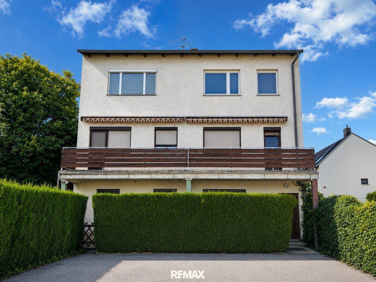 Mehrstöckiges Gebäude mit Balkonen, Rollläden und einer gepflegten Hecke im Vorgarten.