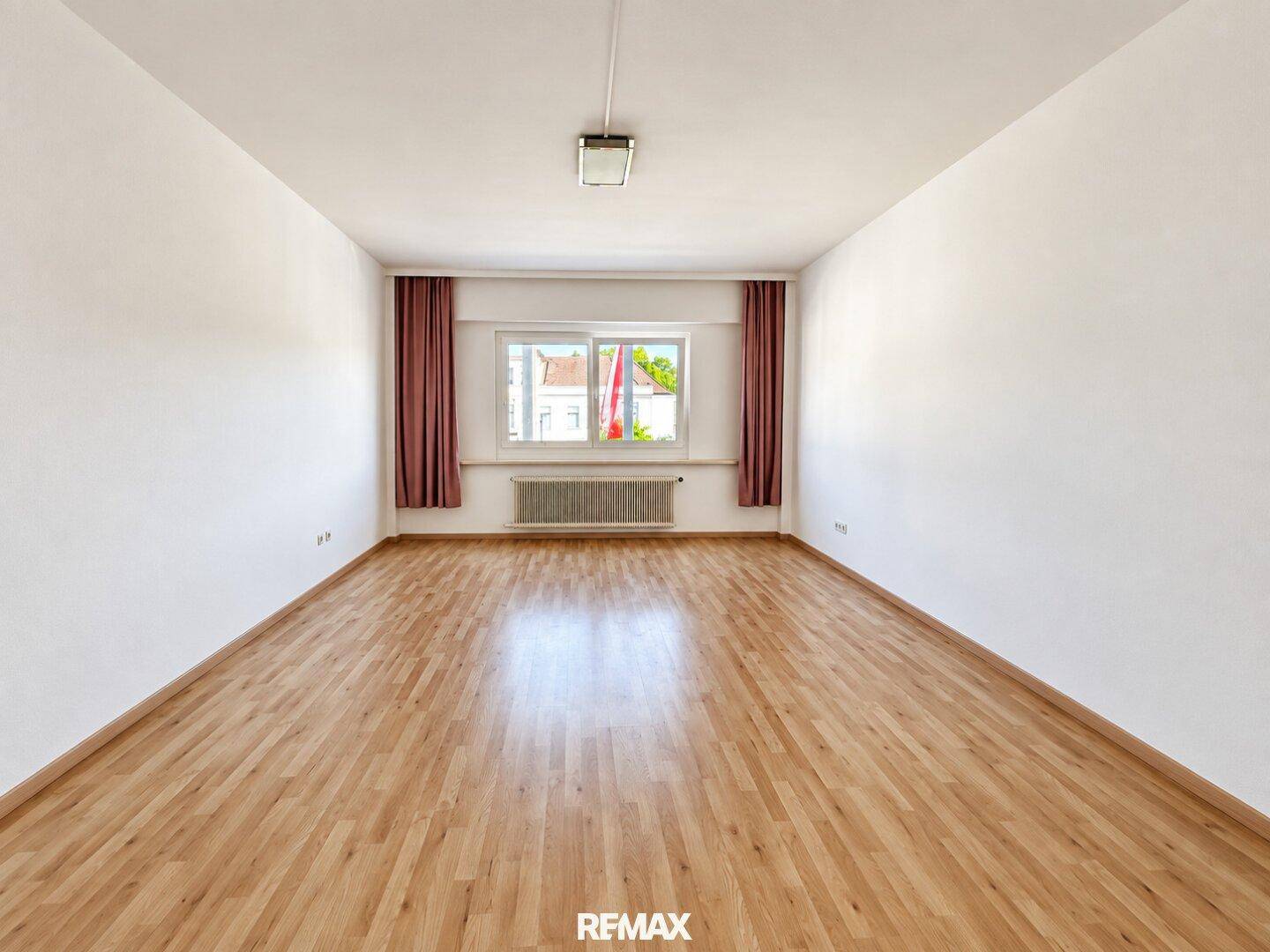 Lichtdurchflutetes Zimmer mit hellem Parkettboden und einem Fenster mit blickdichten Vorhängen.