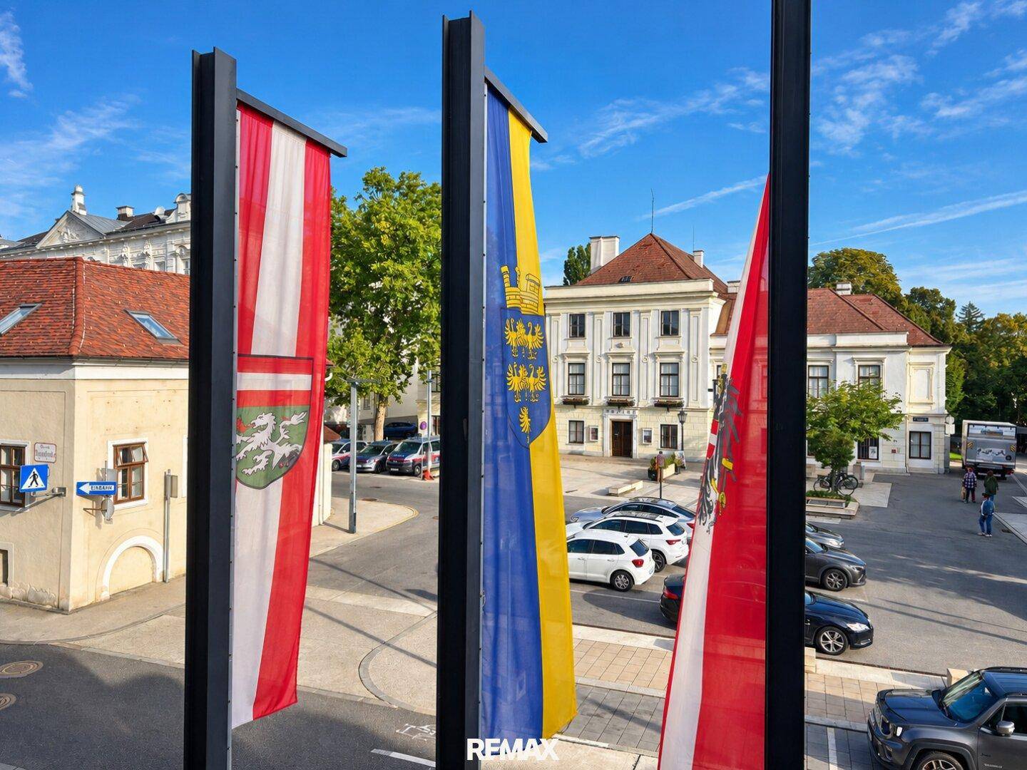Malerischer Stadtplatz mit historischen Gebäuden, parkenden Autos und wehenden österreichischen Flaggen.