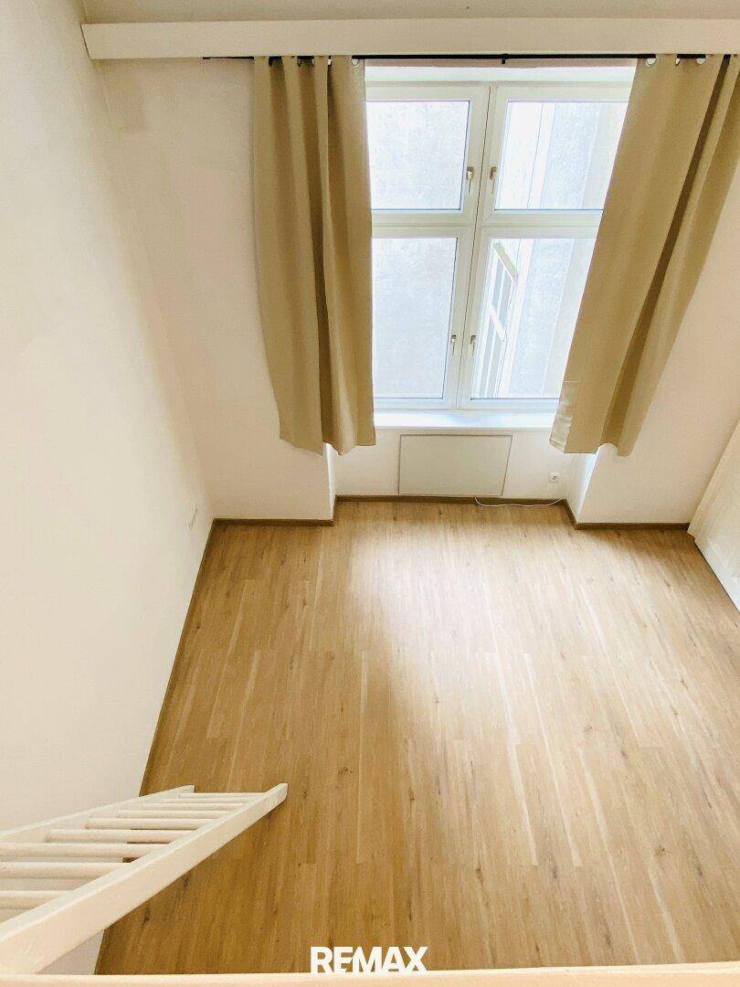 Blick vom Hochbett auf den hellen Holzfußboden und das Fenster mit Vorhängen.