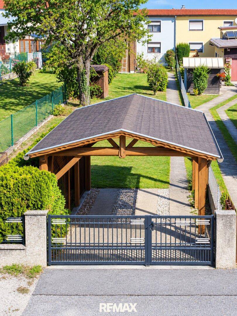 Carport und Zufahrt eines Einfamilienhauses, umgeben von Grünflächen und einem Zaun.