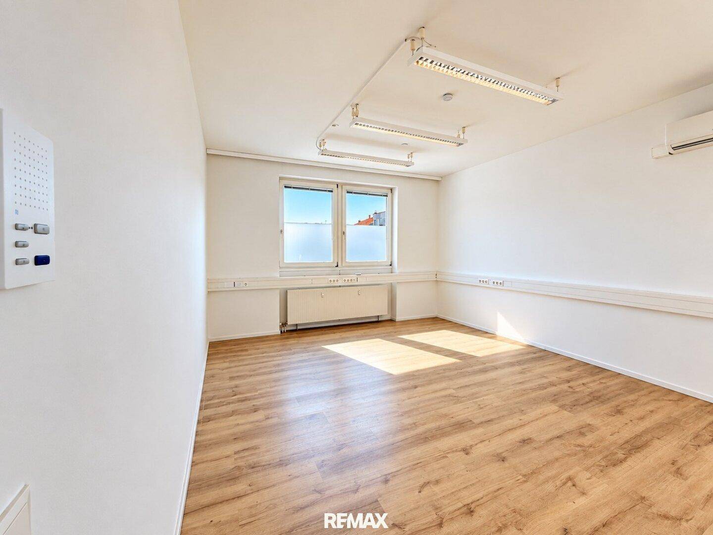 Helles, leeres Büro mit Laminatboden, weißen Wänden und einem Fenster mit Sichtschutzfolie, ausgestattet mit Klimaanlage.