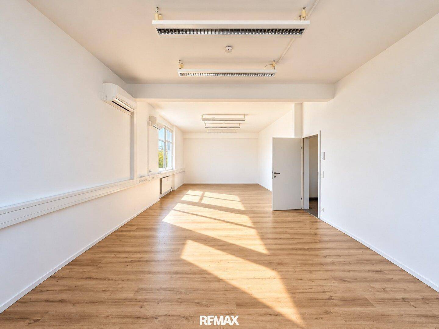 Geräumiges, leeres Büro mit hellem Laminatboden, weißen Wänden und großen Fenstern, die viel Tageslicht hereinlassen.