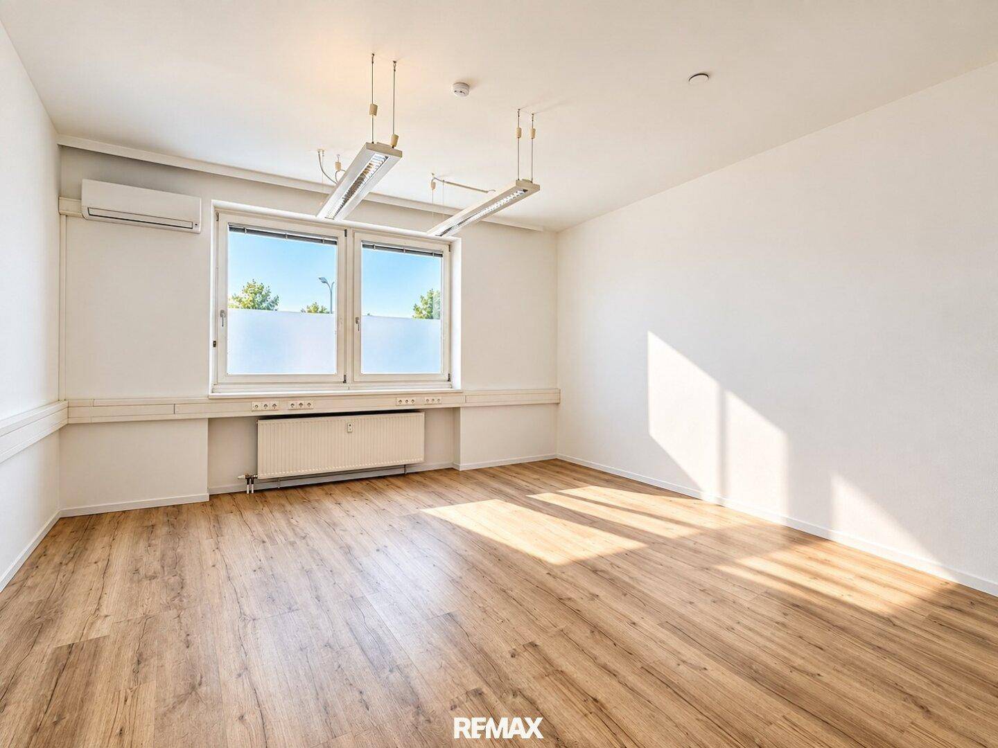 Leeres Büro mit hellem Laminatboden, weißen Wänden und einem Fenster, das für natürliche Belichtung sorgt.
