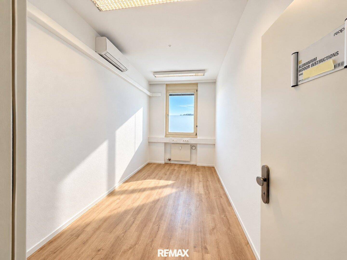 Kleines, helles Büro mit Laminatboden, weißen Wänden, einem Fenster und einer Klimaanlage.