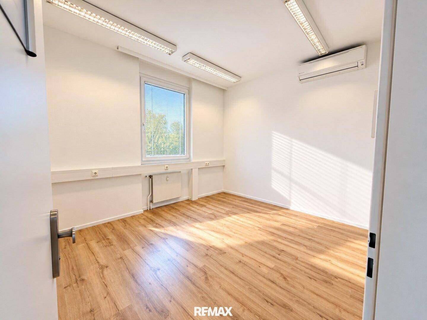Leeres Büro mit Laminatboden, weißen Wänden, einem Fenster mit Jalousien und einer Klimaanlage.