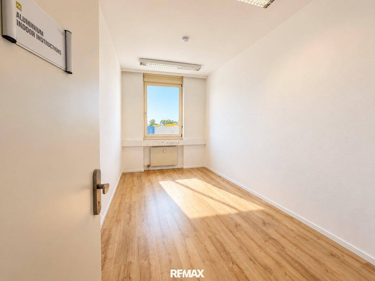 Helles, leeres Büro mit Laminatboden, weißen Wänden und einem Fenster, das viel Sonnenlicht hereinlässt.