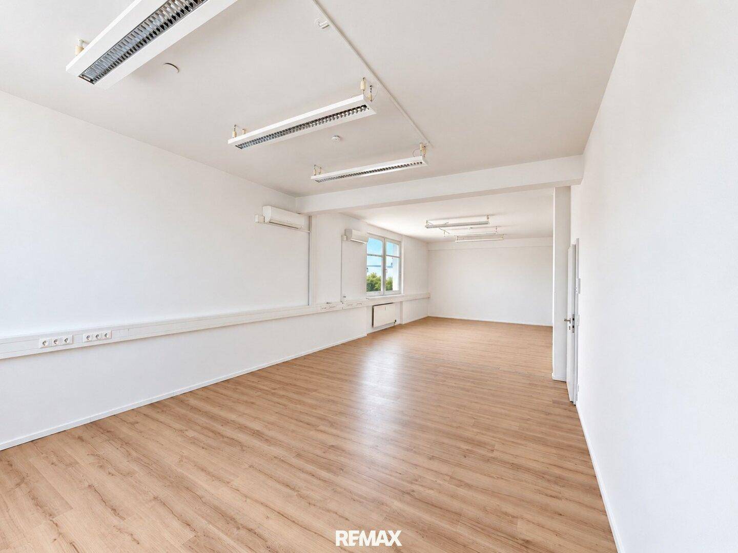 Geräumiges, leeres Büro mit hellem Laminatboden, weißen Wänden und großen Fenstern, die viel Tageslicht hereinlassen.