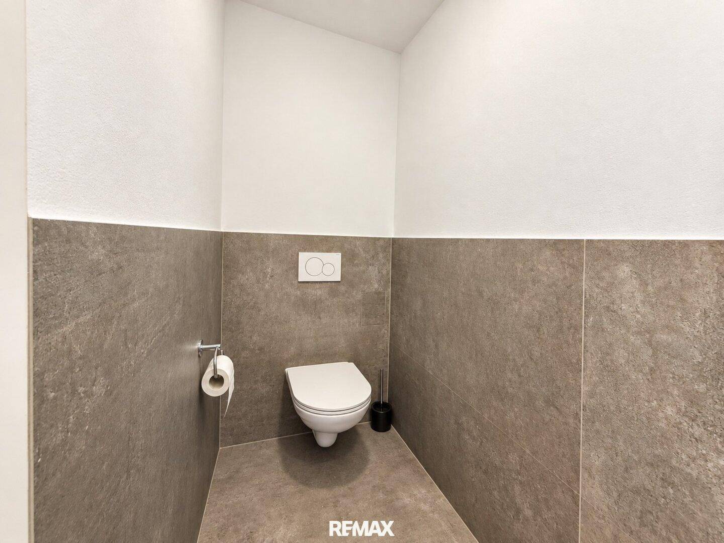 Modernes WC mit wandhängender Toilette und grauen Fliesen an den Wänden, in einem gepflegten Zustand.