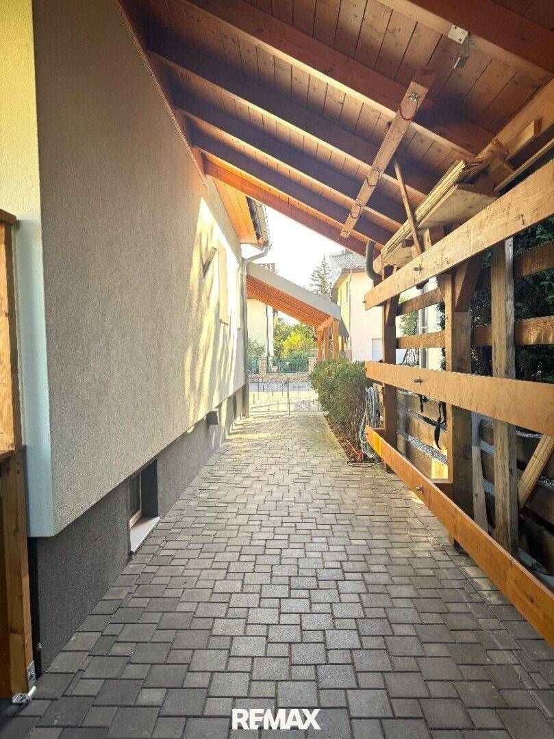 Überdachter Zugangsbereich mit Holzbalkenkonstruktion und gepflastertem Weg neben dem Haus.