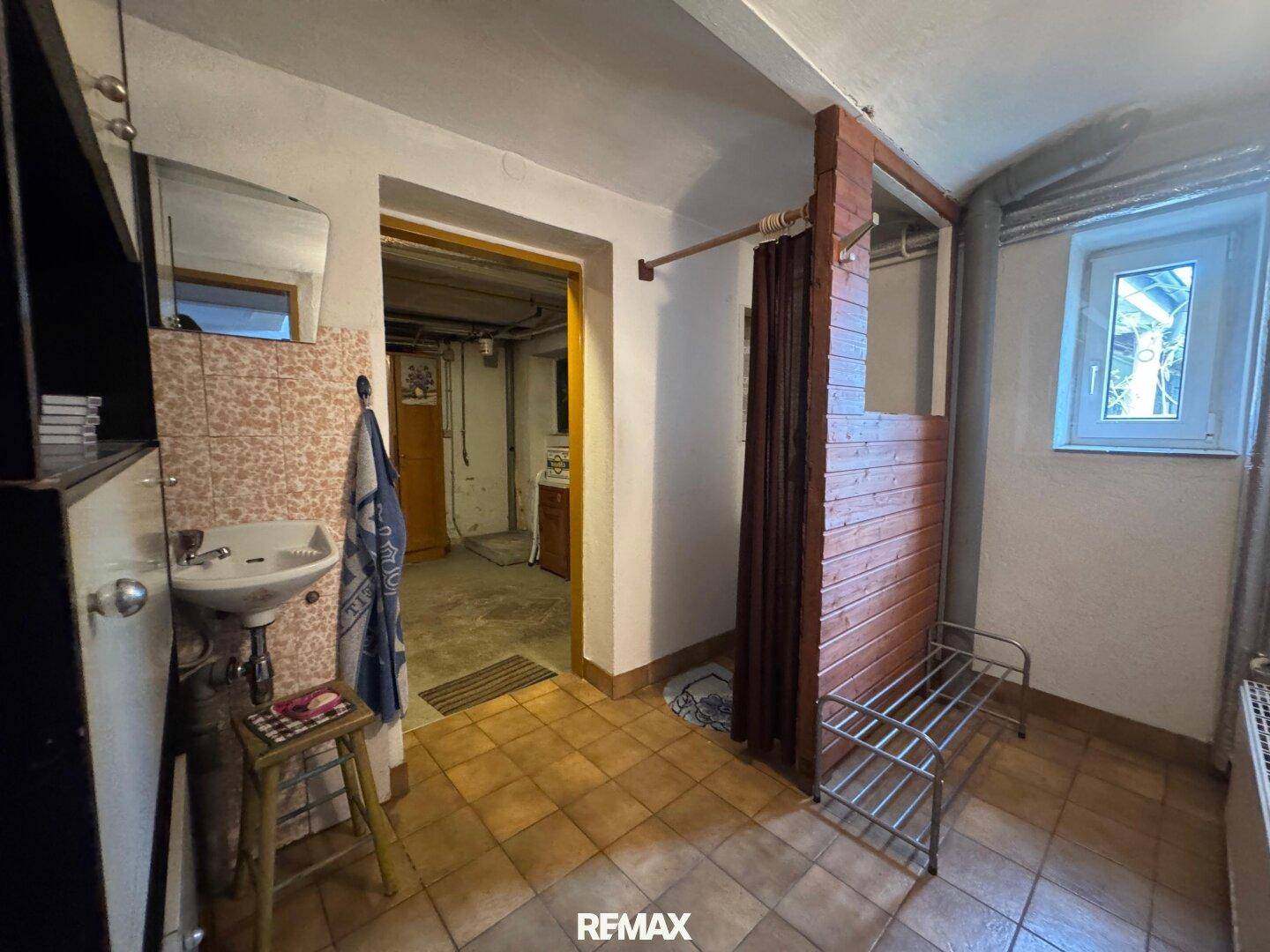 Einfacher Hauswirtschaftsraum im Keller mit Fliesenboden, kleinem Waschbecken und einem Fenster.