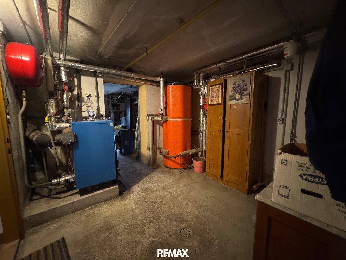 Heizungsraum im Keller mit Boiler, Warmwasserspeicher und sichtbaren Rohren auf Betonboden.