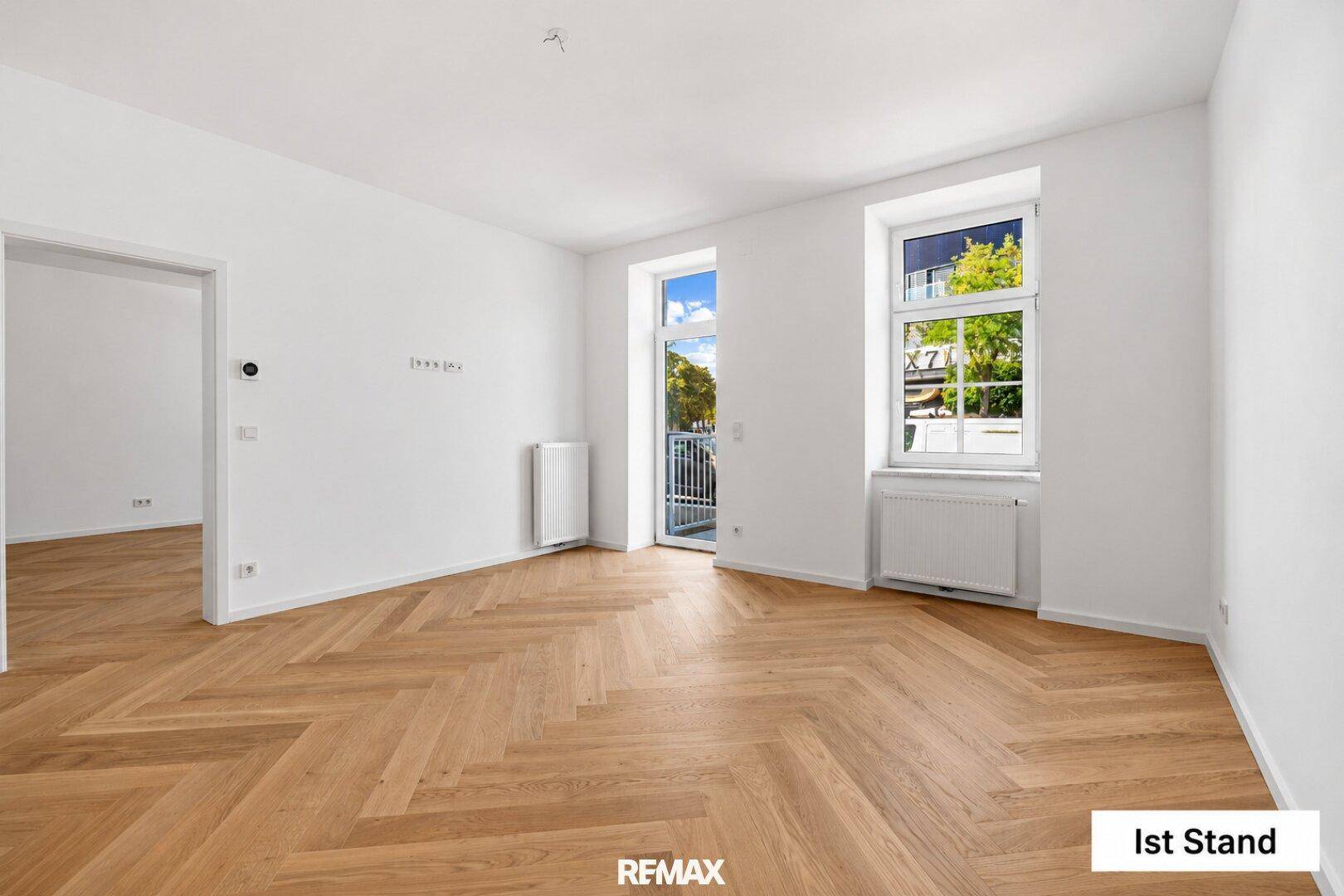 Leeres Wohnzimmer mit Fischgrätparkett, hohen Fenstern und französischem Balkon.