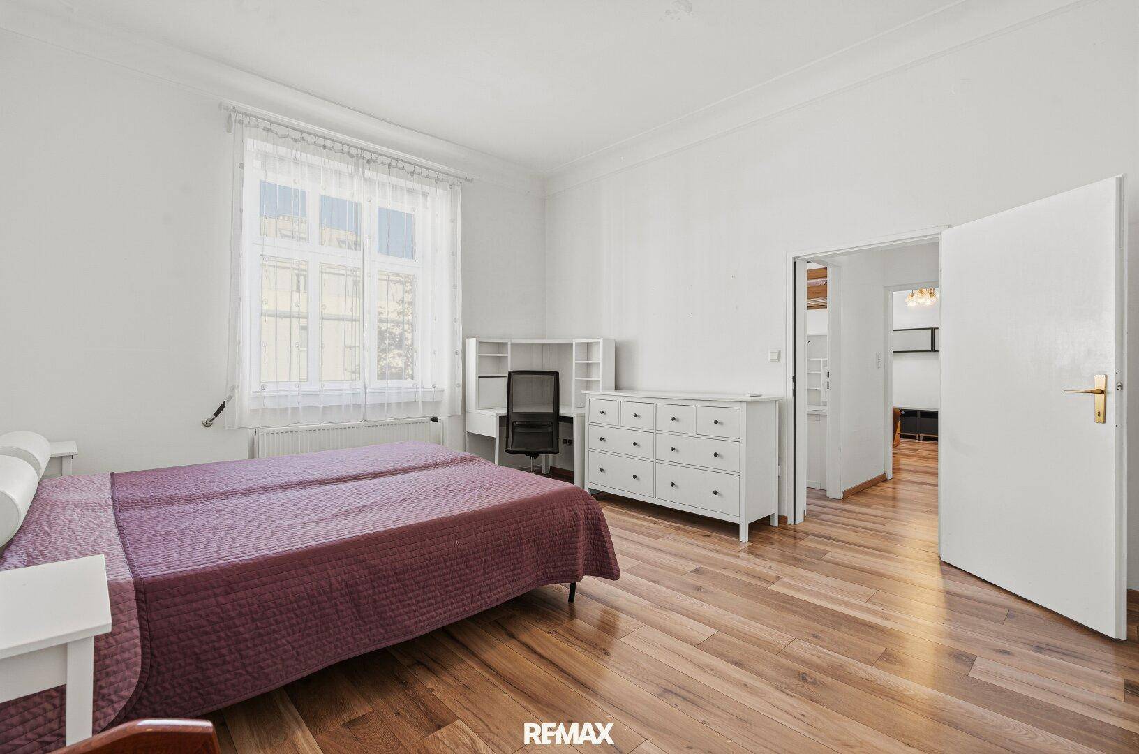 Helles Schlafzimmer mit Holzboden, Doppelbett, Kommode und Zugang zu einem weiteren Raum.