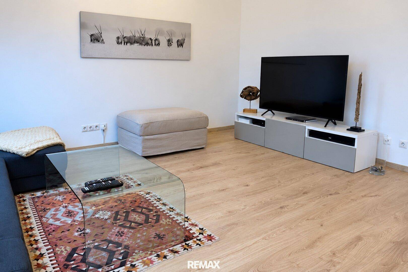 Helles Wohnzimmer mit Fernseher, beigem Ottoman und Holzboden.