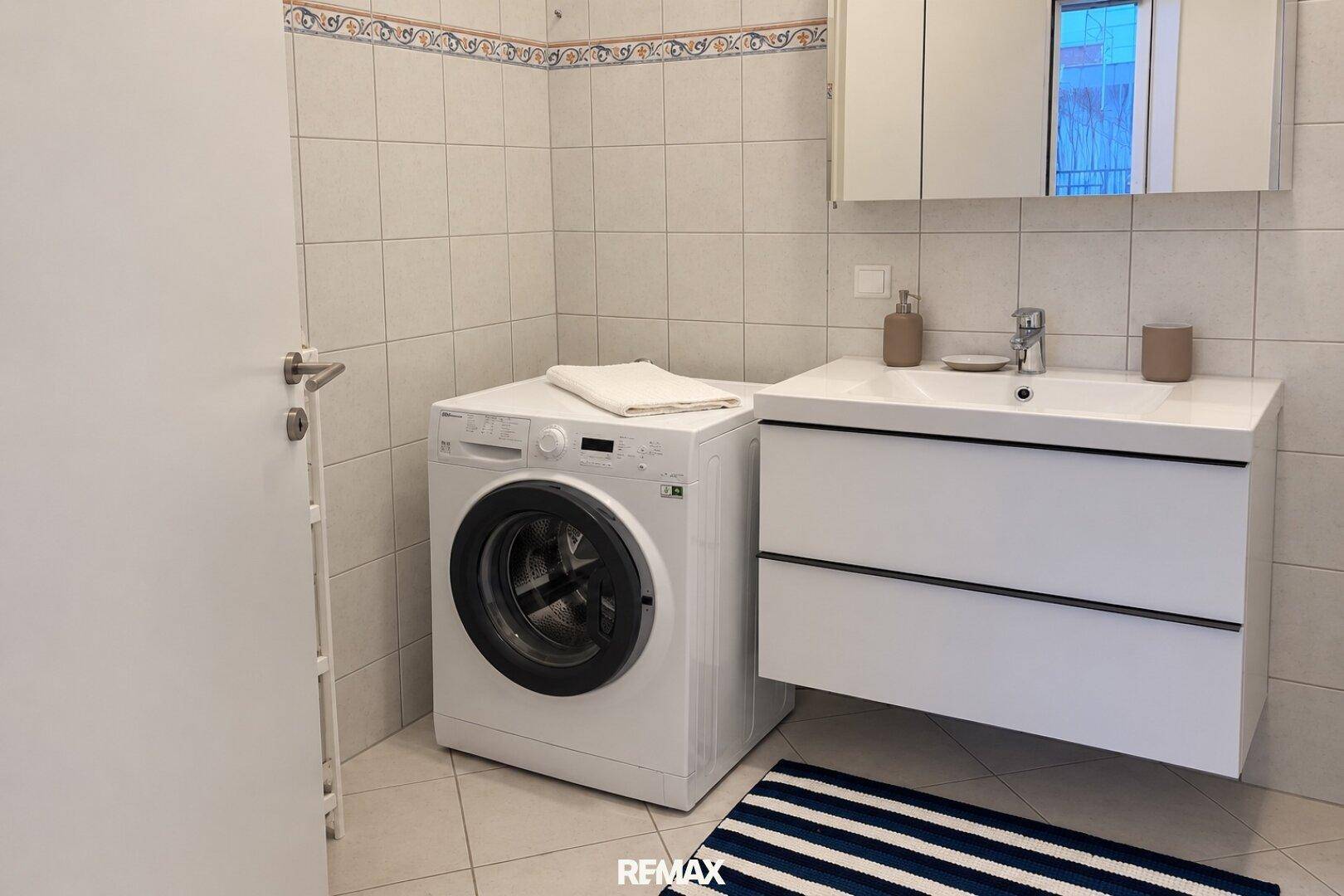 Badezimmer mit Waschmaschine, modernem Waschtisch und hellen Fliesen.