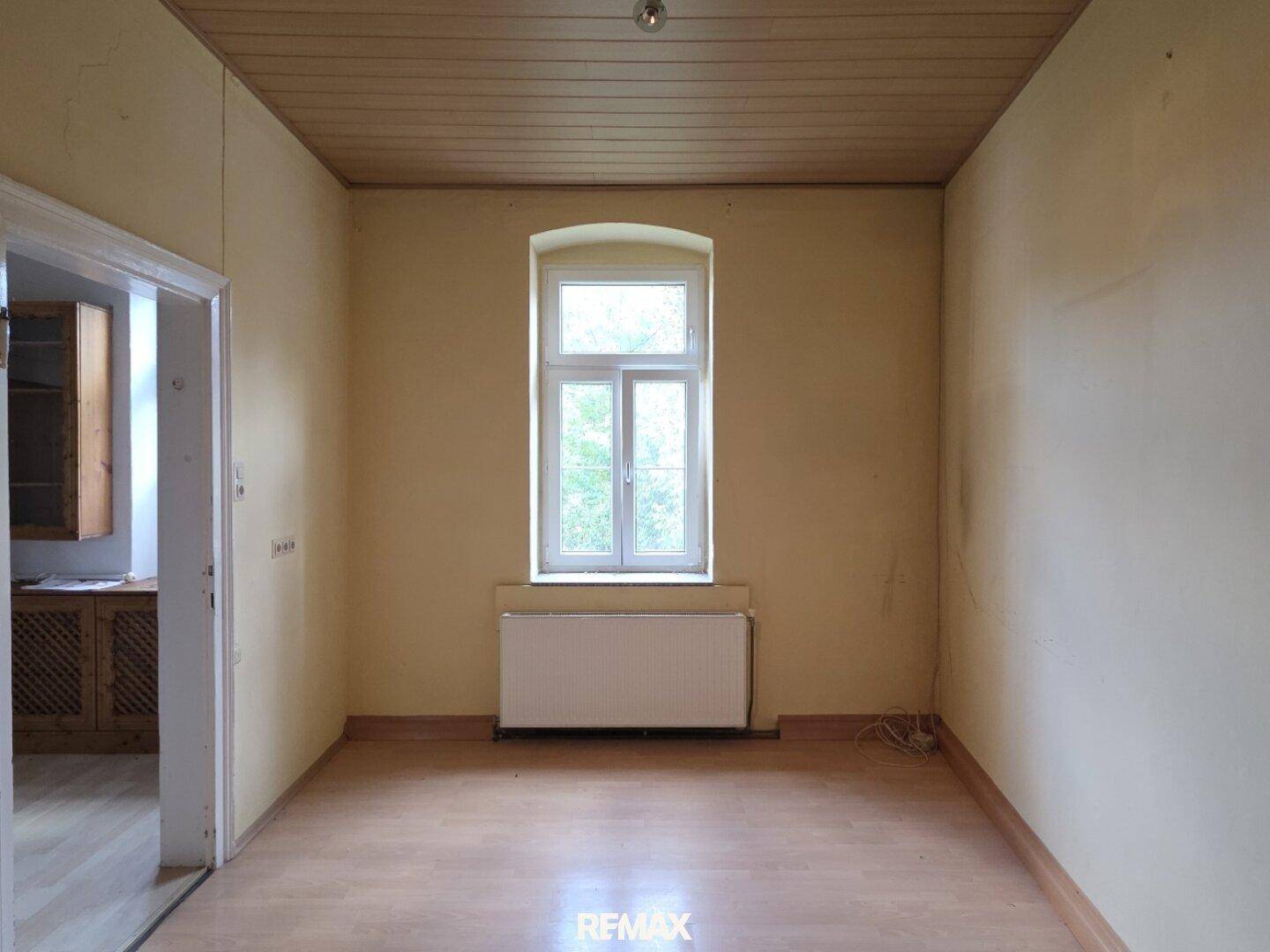 Leerer Raum mit Fenster, Heizkörper und Holzpaneeldecke, bereit für eine individuelle Gestaltung.