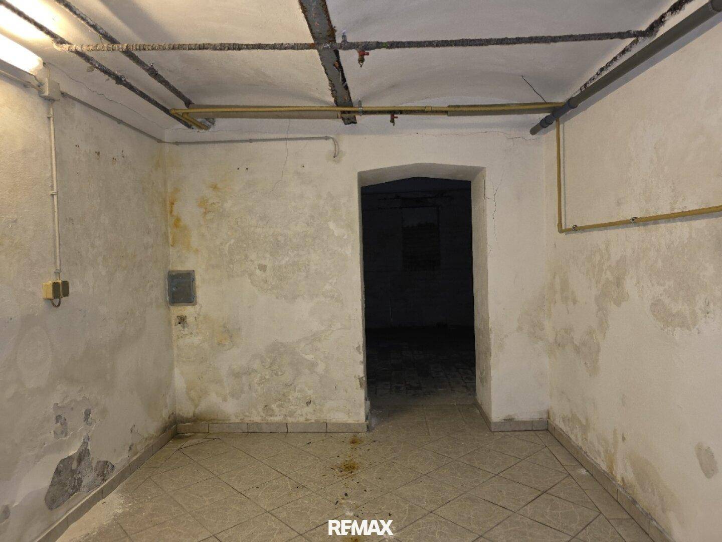 Weiterer Kellerraum mit Fliesenboden, sichtbaren Rohren und einem gewölbten Durchgang zu einem dunkleren Bereich.