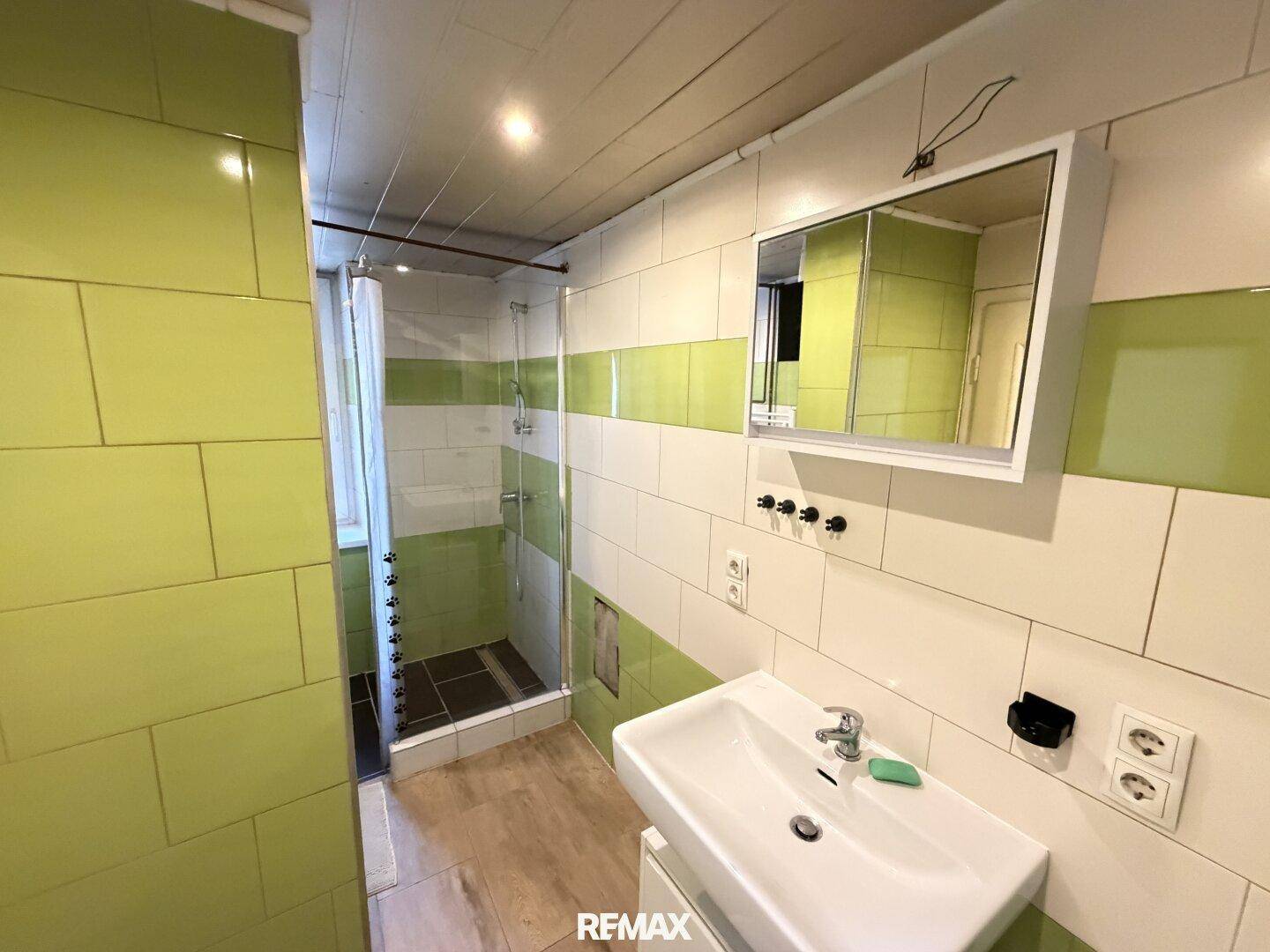 Badezimmer mit grünen und weißen Fliesen, einer Duschkabine und einem einzelnen Waschbecken.