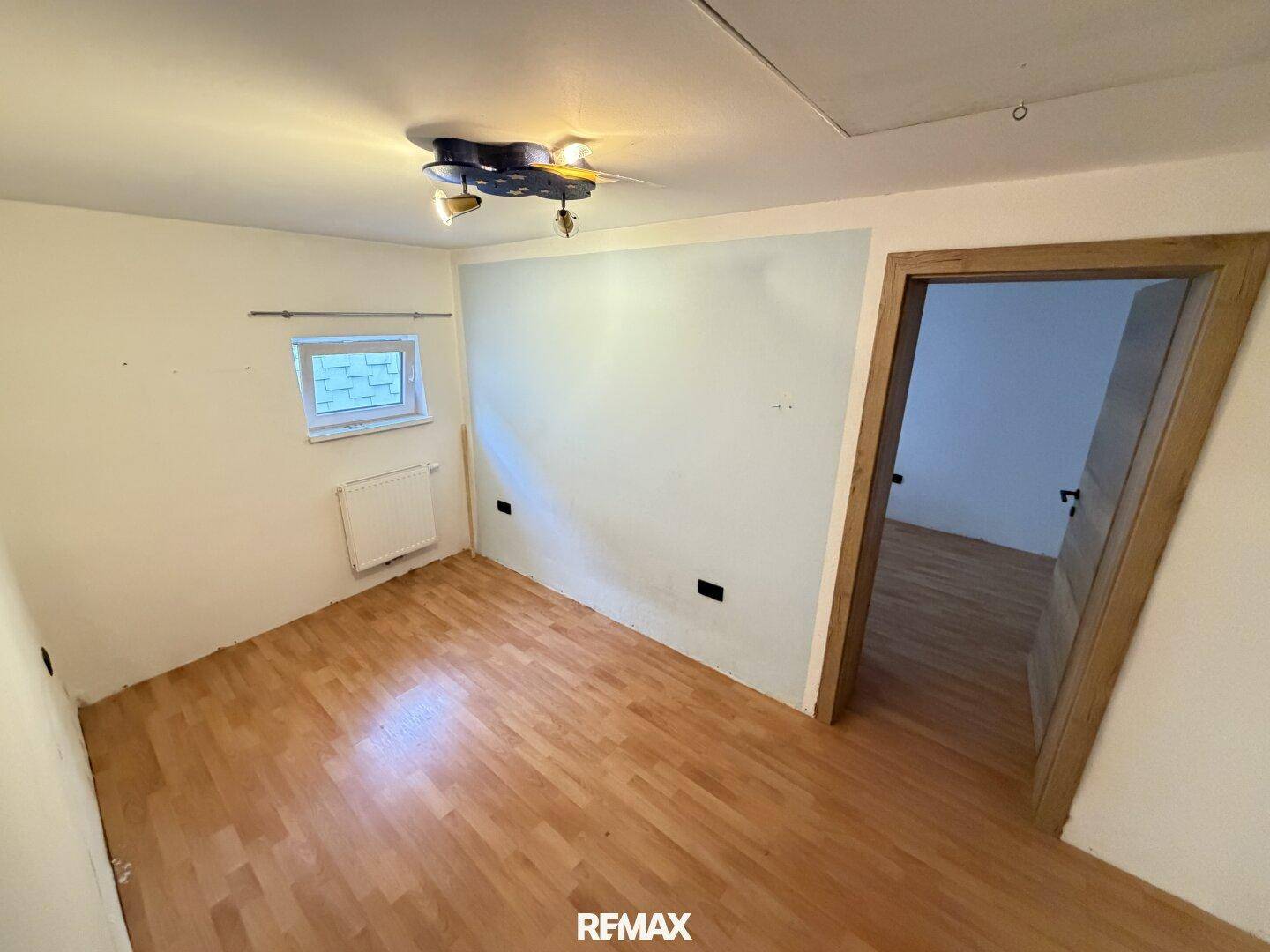 Leeres Zimmer mit hellem Parkettboden, weiß gestrichenen Wänden und einem kleinen Fenster.