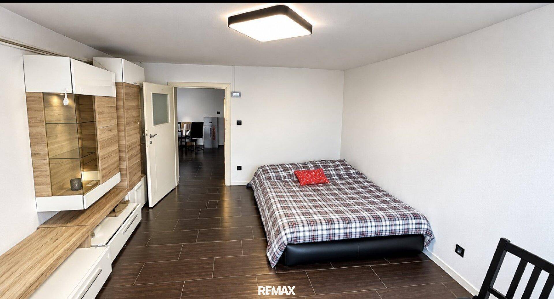 Schlafzimmer mit kariertem Bett, dunklen Fliesen und modernen Möbeln, offen zum Flur.