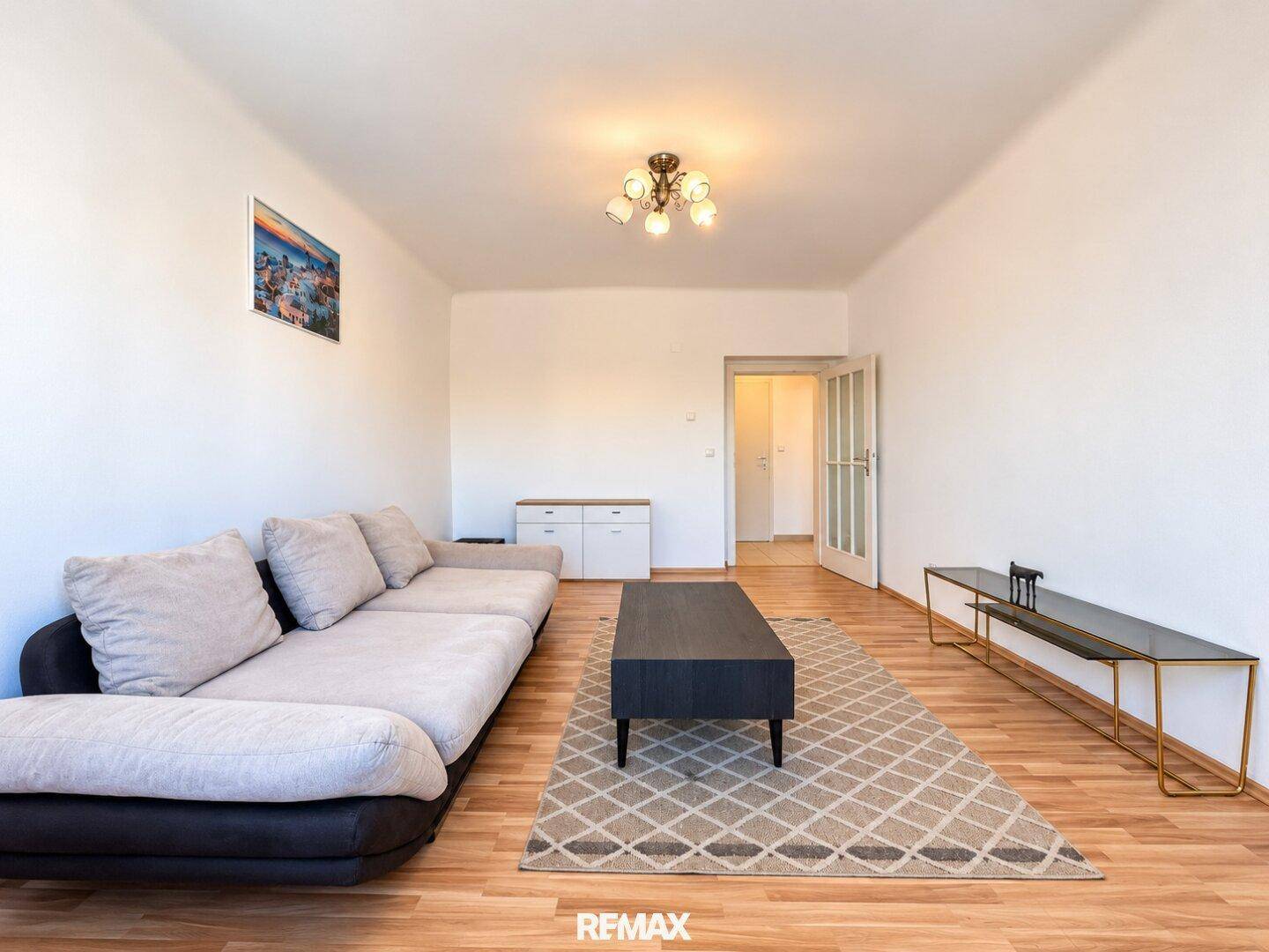 Geräumiges Wohnzimmer mit beigem Sofa, Holzboden und Zugang zu weiteren Räumen.