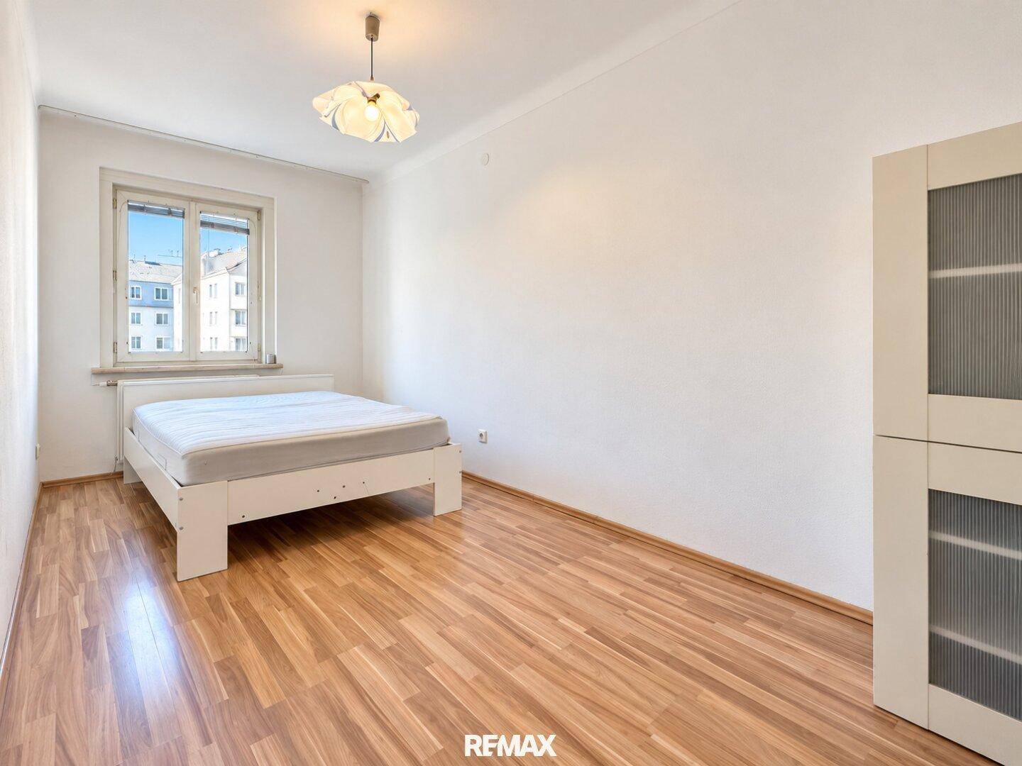 Helles Schlafzimmer mit weißem Bettrahmen, Kleiderschrank und Fenster mit Blick nach draußen.