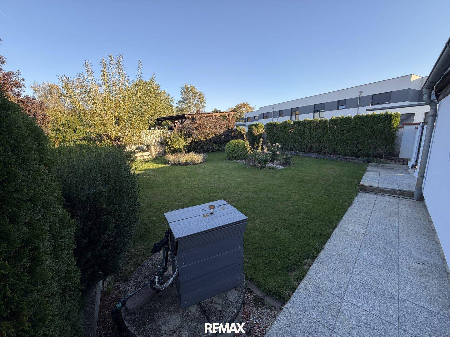 Gepflegter Garten mit Rasenfläche, Hecken und Bäumen unter blauem Himmel.