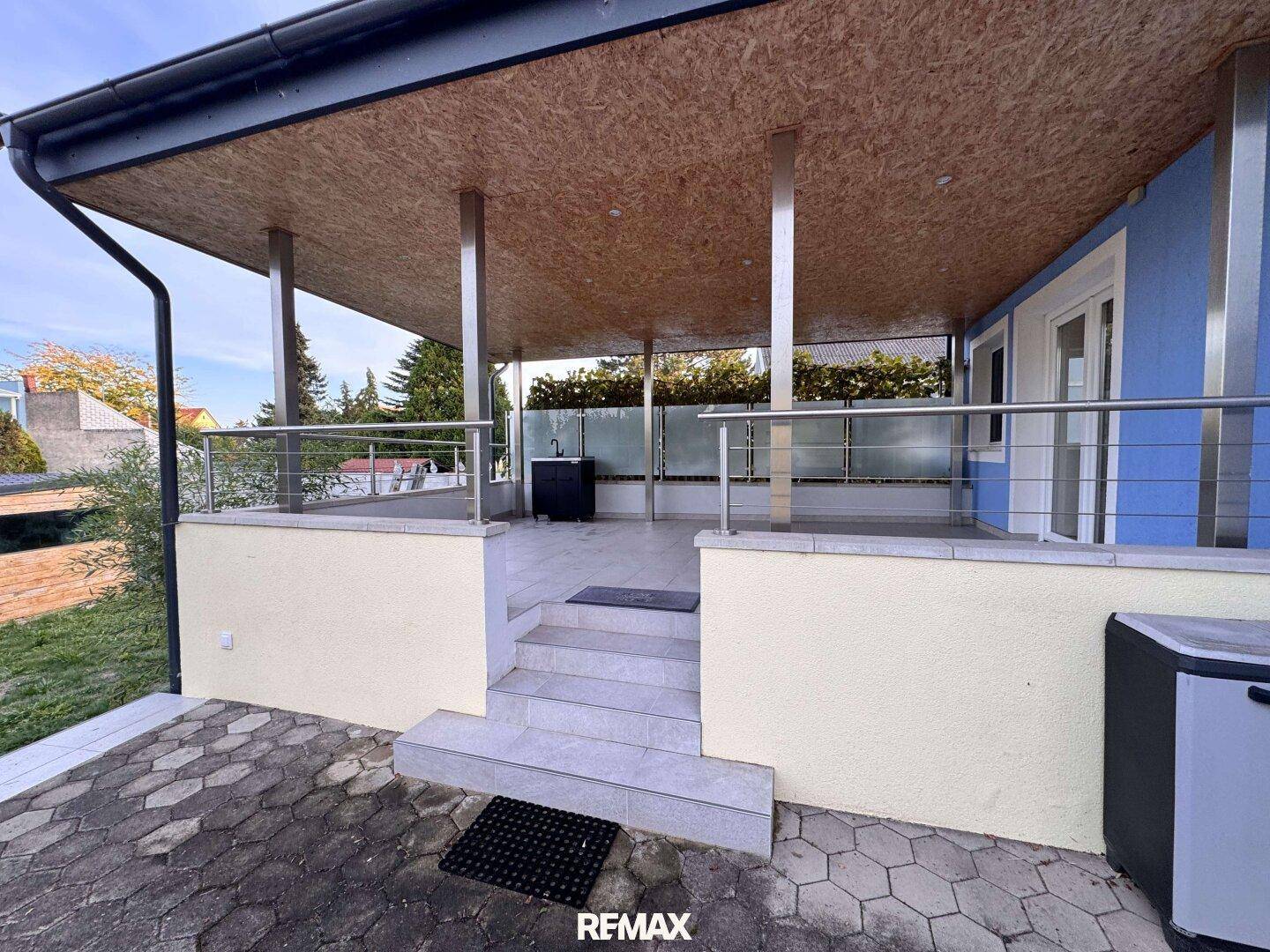 Überdachte Terrasse mit Fliesenboden, Glasgeländer und Blick auf den Gartenbereich.