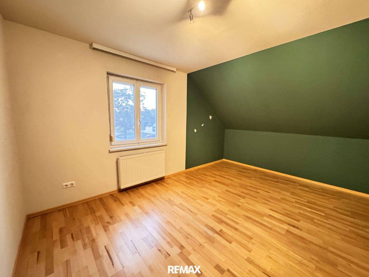 Schlafzimmer mit Holzboden, grüner Akzentwand und Fenster mit Blick nach draußen.