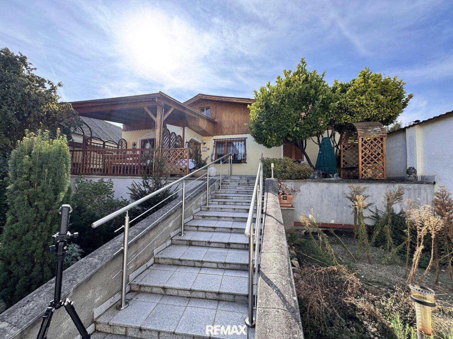 Steintreppe führt zu einem Haus mit großer überdachter Terrasse und Gartenblick.